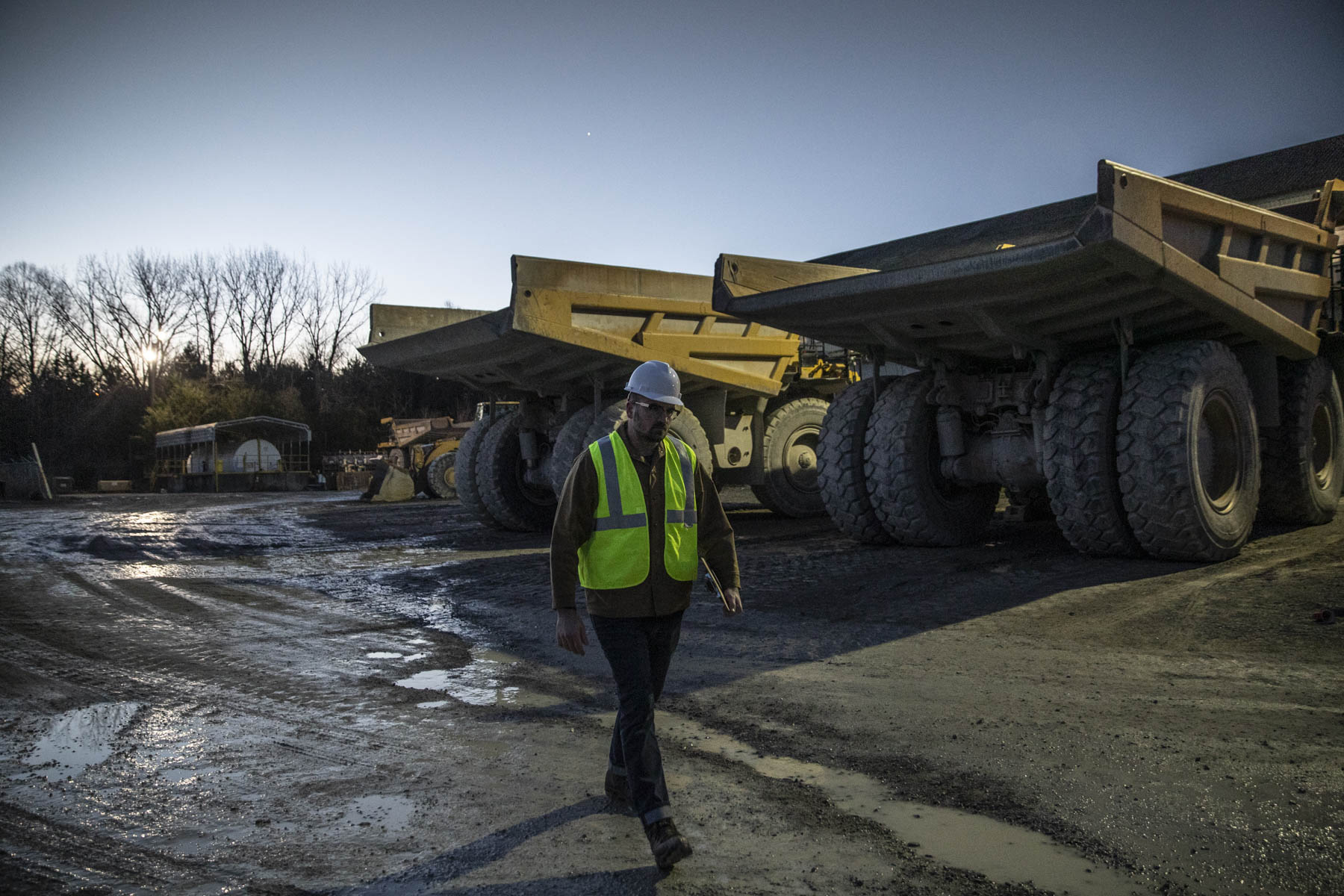 Industrial worker walking toward massive mining haul trucks at dusk