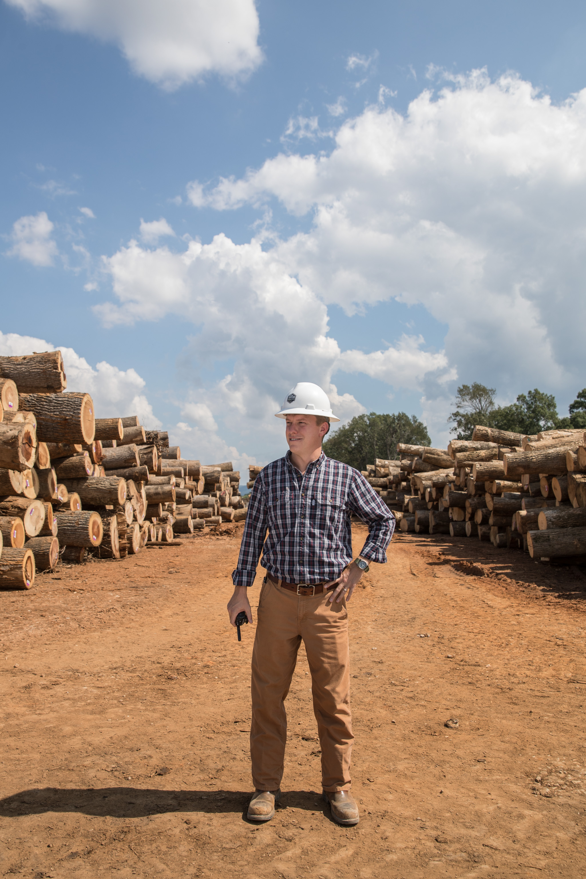 Timber yard worker portrait between stacked logs beneath an open sky