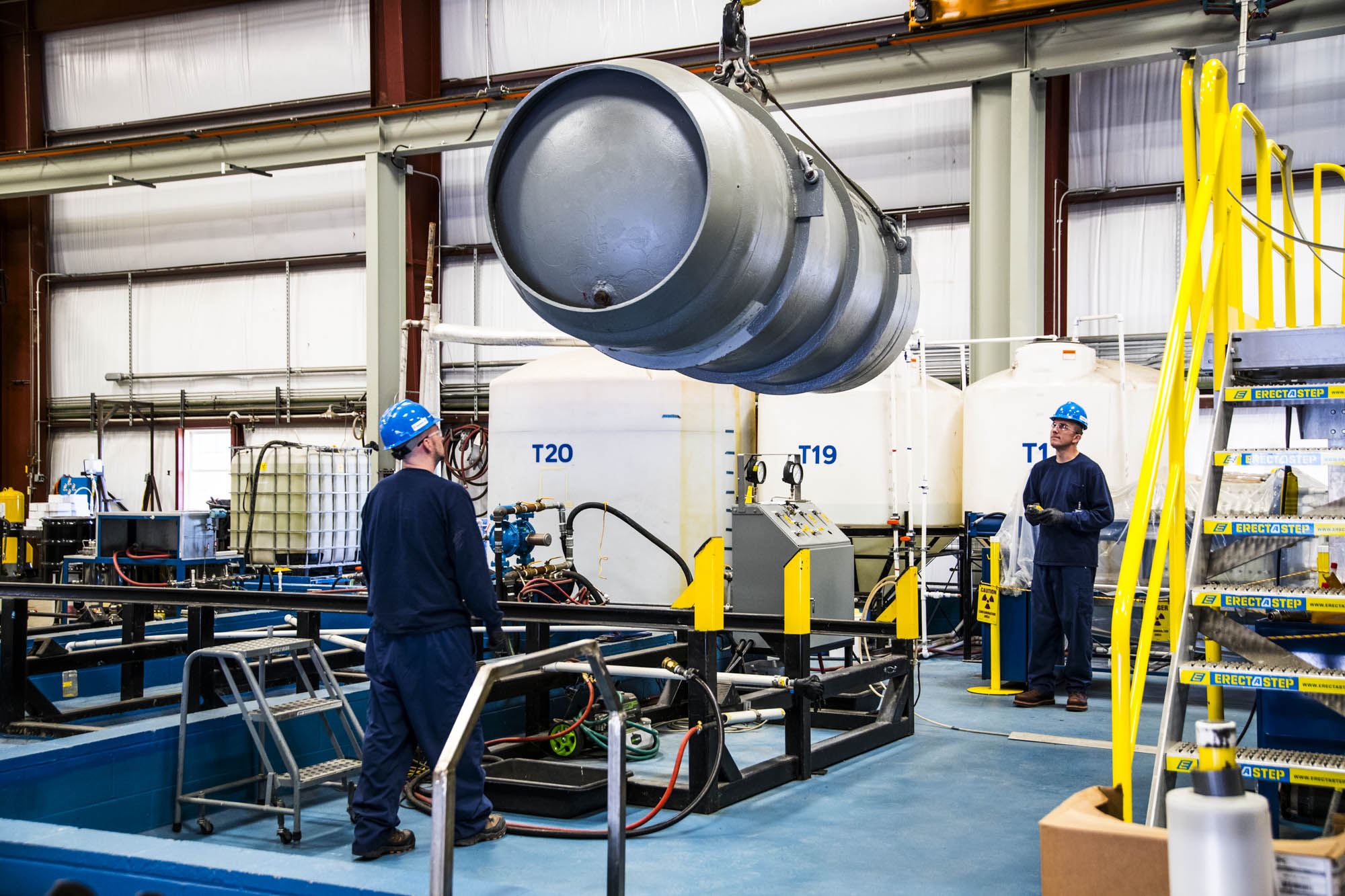 Crane lifting a cask past labeled process tanks inside the facility
