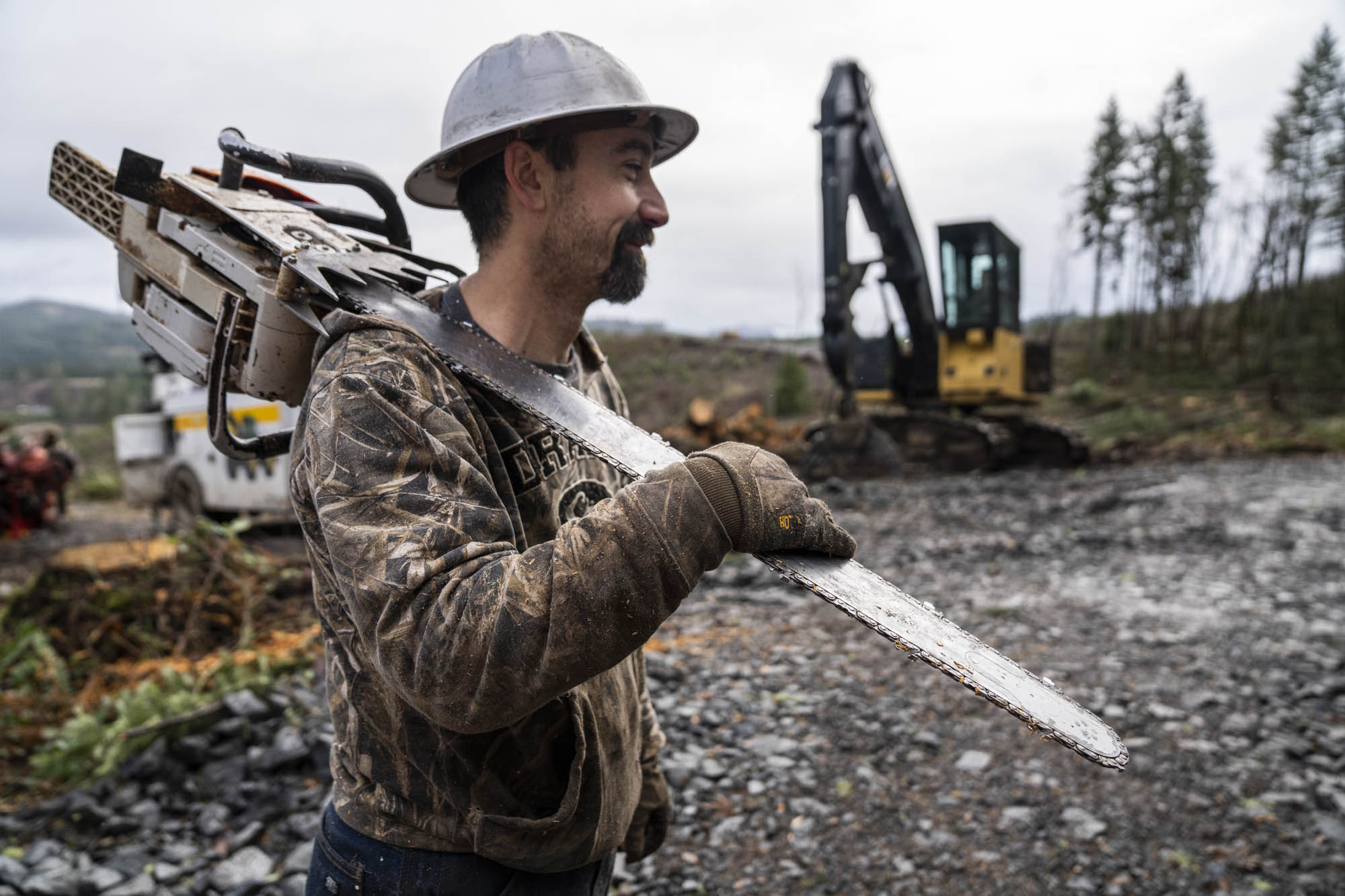 Faller carrying a chainsaw past logging equipment on a cleared hillside